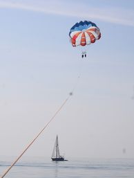 Bright red, white, and blue parasail carrying two riders above a sailboat on calm coastal waters under a pale blue sky.