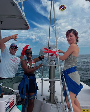Three people on a boat deck in coastal waters wearing parasail harnesses, holding a shark puppet and red claw prop while a colorful parasail floats above the open ocean under a partly cloudy blue sky.