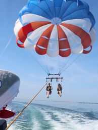 Two people parasailing over a calm coastal ocean under a red, white, and blue parachute towed by a boat with a playful shark head prop, blue sky and wake in view.