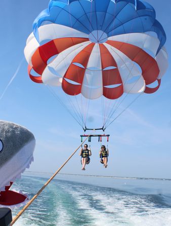 Two people parasailing over a calm coastal ocean under a red, white, and blue parachute towed by a boat with a playful shark head prop, blue sky and wake in view.
