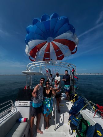 Group of passengers giving thumbs-up on a parasailing boat beneath a red, white and blue parachute canopy over calm coastal waters on a sunny day.