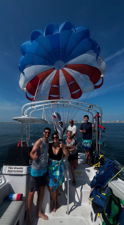 Group of passengers giving thumbs-up on a parasailing boat beneath a red, white and blue parachute canopy over calm coastal waters on a sunny day.