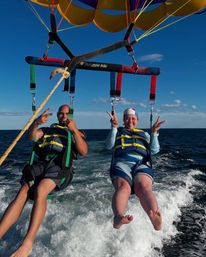 Two parasail riders in life jackets flashing peace signs while tandem parasailing over deep-blue ocean with a yellow parachute canopy and boat wake below