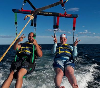 Two parasail riders in life jackets flashing peace signs while tandem parasailing over deep-blue ocean with a yellow parachute canopy and boat wake below