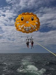 Two people parasailing beneath a giant yellow smiling parasail over the ocean, towed by a boat with its wake under a partly cloudy sky.