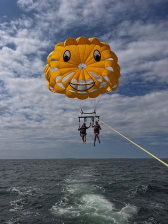 Two people parasailing beneath a giant yellow smiling parasail over the ocean, towed by a boat with its wake under a partly cloudy sky.