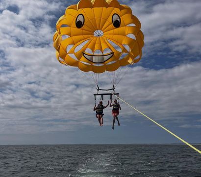 Two people parasailing beneath a giant yellow smiling parasail over the ocean, towed by a boat with its wake under a partly cloudy sky.