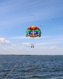 Two people parasailing over a deep-blue ocean on a sunny day, suspended from a colorful rainbow parachute with a distant coastline below.