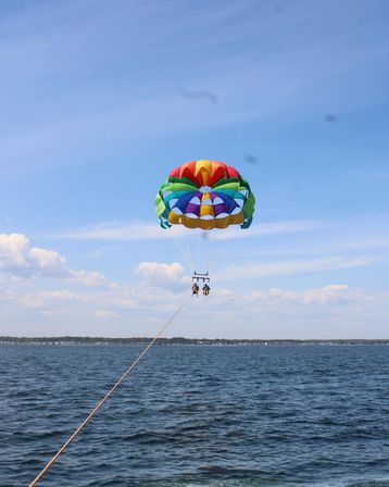 Two people parasailing over a deep-blue ocean on a sunny day, suspended from a colorful rainbow parachute with a distant coastline below.
