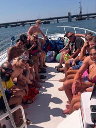 Smiling group enjoying a crowded motorboat cruise on coastal waters near a bridge on a sunny summer day, wearing swimwear and surrounded by bags and gear.