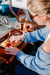 Sunlit scene of a person in a blue sweater preparing a cheese and charcuterie board with sliced cheese, cured meats, a wine glass and beer bottle on a sailboat cockpit.