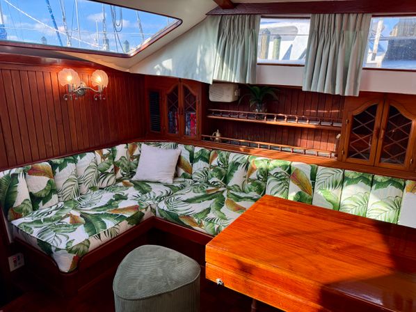 Cozy yacht salon interior with varnished wood paneling, L-shaped banquette upholstered in tropical leaf fabric, wooden dining table, porthole windows and soft curtains with a dockside marina view.