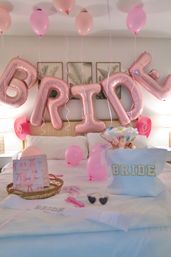 Bridal suite bachelorette setup with rose-gold foil letter balloons spelling 'BRIDE', pink latex balloons floating over a hotel-style bed, a 'BRIDE' tote, gift bag and accessories on white linens