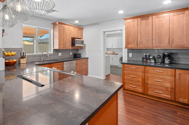 Bright modern kitchen with warm wood cabinets, dark quartz island and countertops, stainless sink and appliances, pendant lights, and view into adjacent laundry room and backyard.