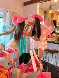 Two women in pink cowgirl hats sipping orange cocktails at a bright indoor kitchen party with pink gift bags, inflatable cactus decor and citrus slices