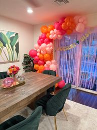 Indoor dining room party scene with a pink and orange balloon garland draped over a metallic fringe curtain and gold letter banner, rustic wooden table topped with disco balls and a bright floral centerpiece, emerald velvet chairs and tropical wall art.