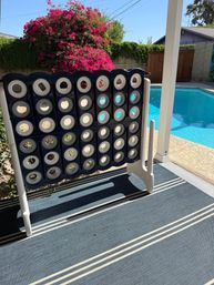Oversized blue-and-white Connect Four game panel on a striped patio rug beside a bright blue backyard pool, with vibrant pink bougainvillea and green hedge under a sunny sky.