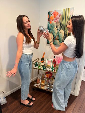 Two smiling women clinking mirrored disco-ball tumblers in a home living space beside colorful cactus wall art and a gold bar cart stocked with champagne and party decor.