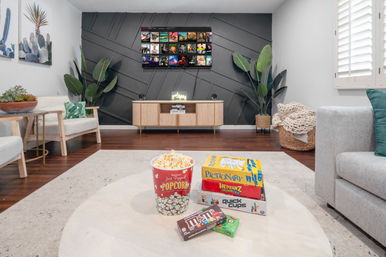 Modern cozy living room entertainment setup with wall-mounted TV, textured dark accent wall, wooden media console and tall potted plants; coffee table in foreground with popcorn tub, candy and stacked board games ready for family game night.