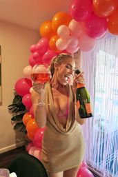Woman celebrating indoors with a champagne bottle and large rosé glass in front of a pink and orange balloon garland and fringe party backdrop
