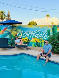 Two people relaxing by a turquoise pool in front of a colorful Scottsdale cactus mural — woman on a lounge chair under a blue umbrella and a man sitting at the pool edge with his feet in the water on a sunny day.