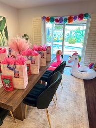 Bright pool-party scene: iridescent gift bags with pink tissue on a wooden table, pampas centerpiece, colorful pom-pom garland over sliding doors, and inflatable flamingo and unicorn by backyard pool.