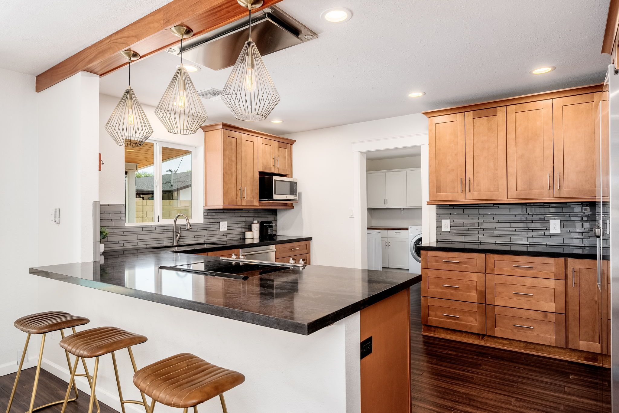 Modern open-plan kitchen with warm wood cabinets, dark stone island and cooktop, three brown leather bar stools, geometric pendant lights, gray subway-tile backsplash, stainless appliances and a view into a laundry room.