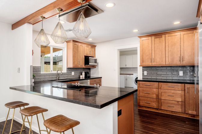 Modern open-plan kitchen with warm wood cabinets, dark stone island and cooktop, three brown leather bar stools, geometric pendant lights, gray subway-tile backsplash, stainless appliances and a view into a laundry room.