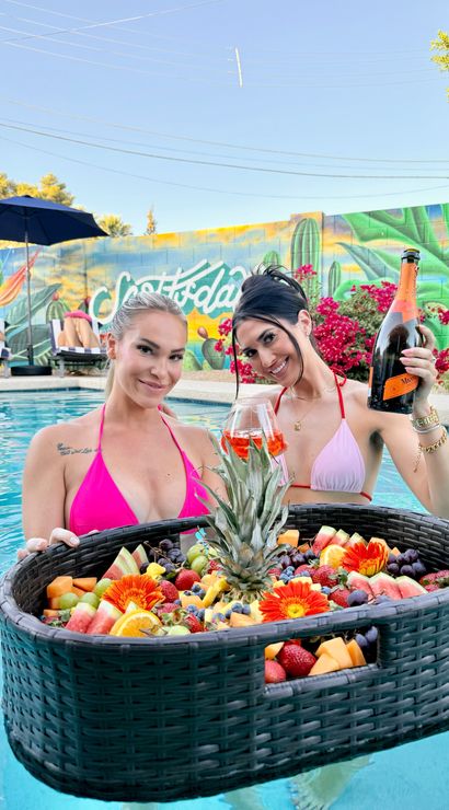 Two women in pink bikinis smiling in a sunny backyard pool, one holding a bottle of champagne and a glass beside a large floating wicker fruit tray with pineapple and flowers, colorful mural and lounge chairs in the background