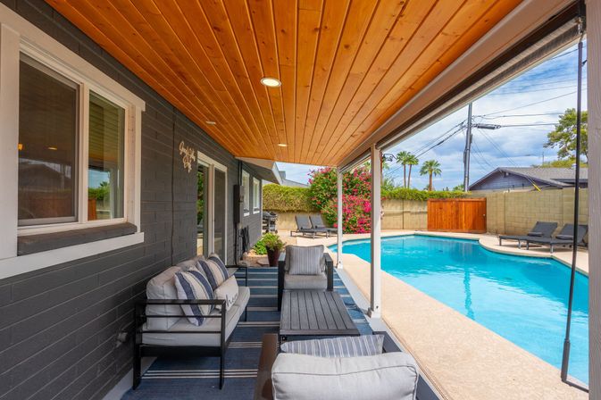 Covered backyard patio with wooden-plank ceiling, cushioned outdoor seating and coffee table beside a curved blue swimming pool with lounge chairs, flowering shrubs and palm trees.