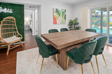 Scottsdale dining area with a chunky reclaimed-wood table, emerald velvet chairs with gold legs, rattan hanging chair by a faux boxwood wall and sliding doors to a pool.