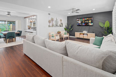 Bright open-concept living and dining room with light-gray sectional, dark geometric accent wall with wall-mounted TV, hardwood floors, mid-century armchairs, green accent pillows and plants, and a dining table by sliding glass doors.