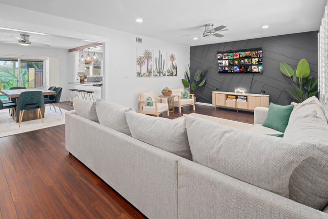 Bright open-concept living and dining room with light-gray sectional, dark geometric accent wall with wall-mounted TV, hardwood floors, mid-century armchairs, green accent pillows and plants, and a dining table by sliding glass doors.