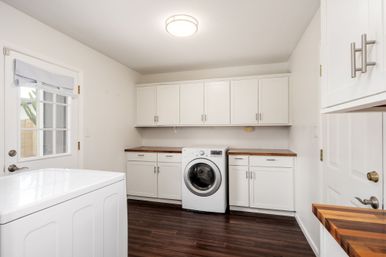 Bright, tidy modern laundry room with front-loading washer flanked by white shaker cabinets, warm wood countertops, dark hardwood floor and a glass-paneled door letting in natural light.