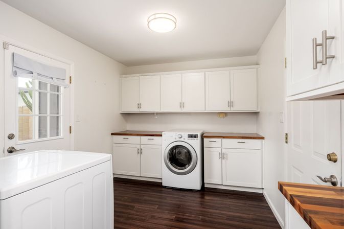 Bright, tidy modern laundry room with front-loading washer flanked by white shaker cabinets, warm wood countertops, dark hardwood floor and a glass-paneled door letting in natural light.