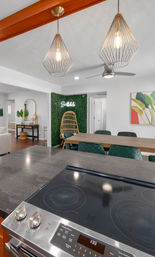 Open-concept Scottsdale kitchen and dining area — dark island with glass cooktop, wooden dining table with green quilted chairs, geometric wire pendant lights, rattan accent chair against a faux-plant wall with neon Scottsdale sign.