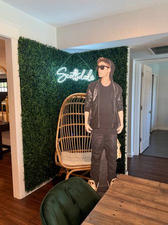 Neon "Scottsdale" sign on a green faux hedge backdrop with a rattan egg chair and a life‑size cardboard cutout of a young man in a black hoodie and sunglasses in a modern indoor dining area.
