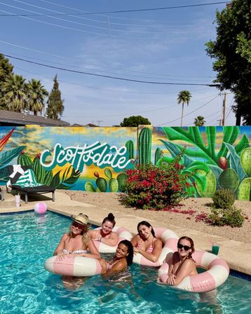 Five friends lounging on pink-and-white inflatable rings in a backyard pool in Scottsdale, Arizona, with a colorful cactus mural reading 'Scottsdale', palm trees and a bright blue sky.
