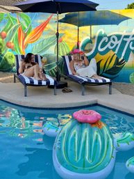 Two women relaxing on striped chaise lounges by a backyard swimming pool, toasting drinks under a patio umbrella in front of a colorful tropical mural, with a bright inflatable float in the water.