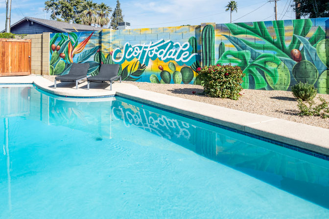 Sunny Scottsdale backyard pool with two black lounge chairs and a vibrant cactus and succulent mural reflected in clear turquoise water.