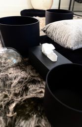 Close-up of a modern home decor vignette: matte black cylindrical planters and a black tissue box on a gray faux-fur rug, with a silver velvet pillow and glass cloche for a cozy, minimalist living room look.