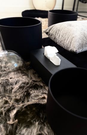 Close-up of a modern home decor vignette: matte black cylindrical planters and a black tissue box on a gray faux-fur rug, with a silver velvet pillow and glass cloche for a cozy, minimalist living room look.