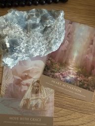 Pale blue crystal cluster resting on pink floral oracle cards reading “Heart Activation” and “Move With Grace” on a wooden tabletop — spiritual crystal healing, self-love meditation setup.