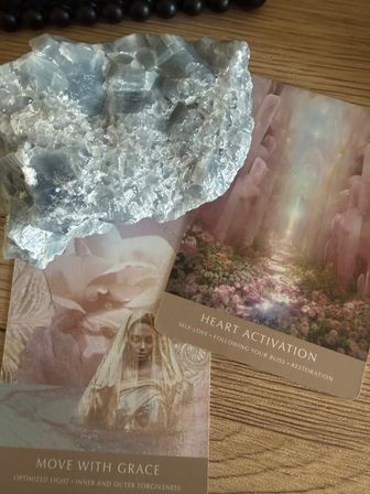 Pale blue crystal cluster resting on pink floral oracle cards reading “Heart Activation” and “Move With Grace” on a wooden tabletop — spiritual crystal healing, self-love meditation setup.