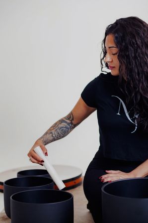 Person with curly hair and tattooed forearm striking black singing bowls with a white mallet in a calming sound‑healing meditation session.