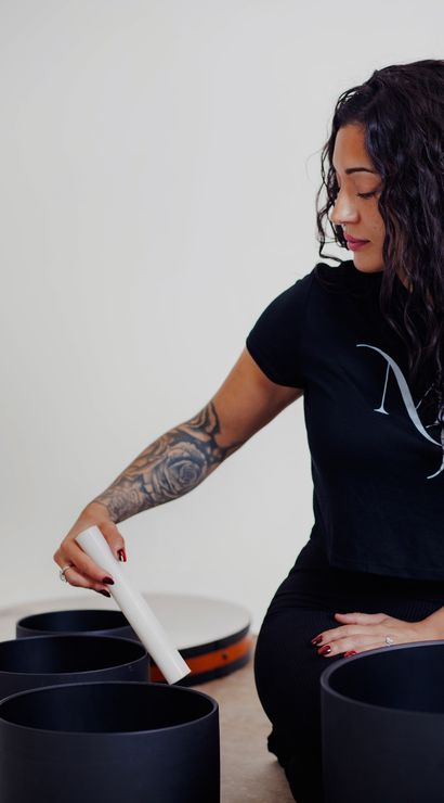 Person with curly hair and tattooed forearm striking black singing bowls with a white mallet in a calming sound‑healing meditation session.