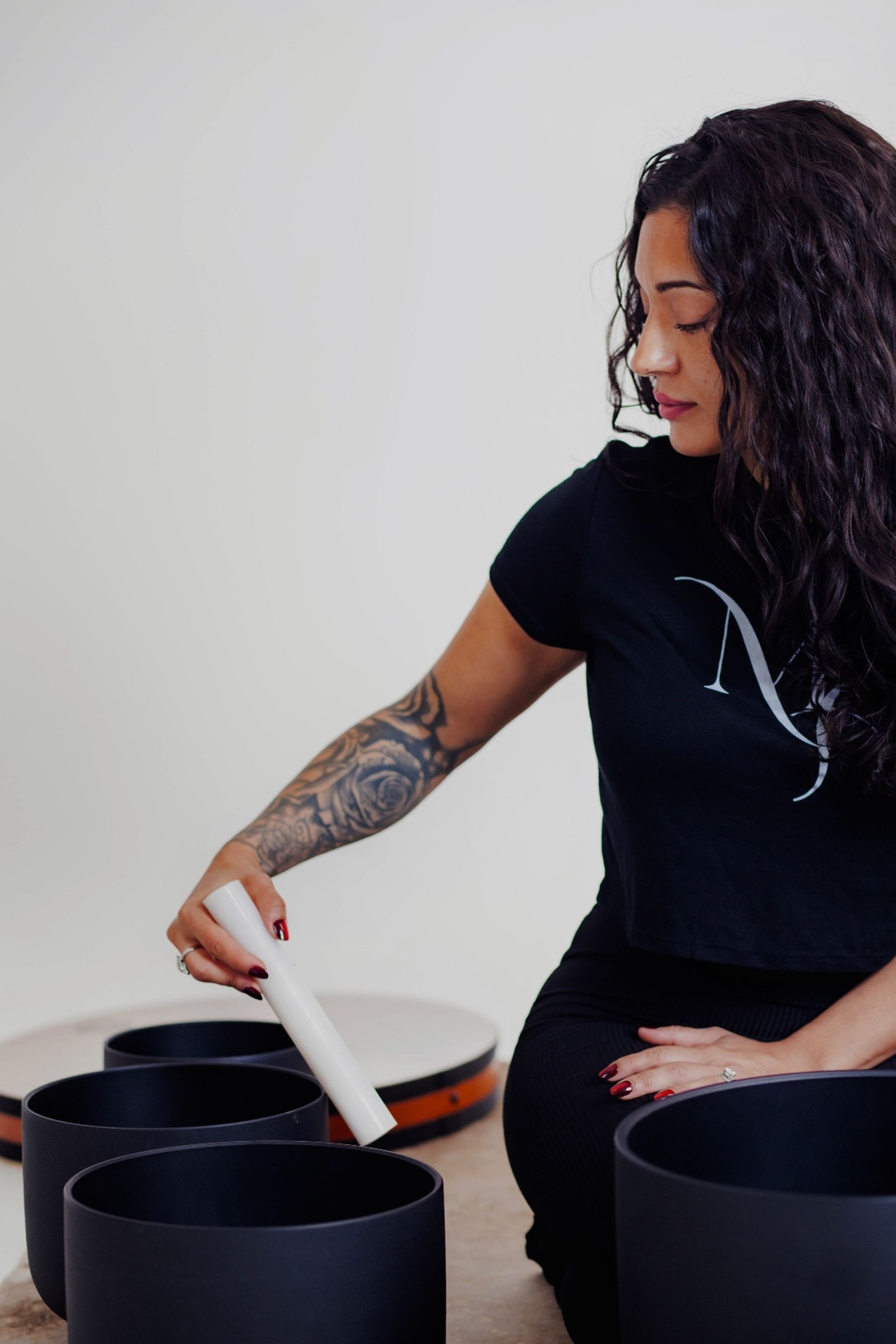Person with curly hair and tattooed forearm striking black singing bowls with a white mallet in a calming sound‑healing meditation session.