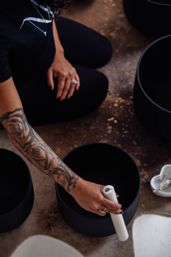 A tattooed arm kneeling on a studio floor strikes a black singing bowl with a white mallet during a sound-bath meditation.