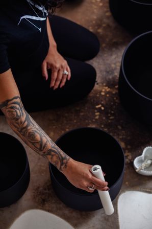 A tattooed arm kneeling on a studio floor strikes a black singing bowl with a white mallet during a sound-bath meditation.