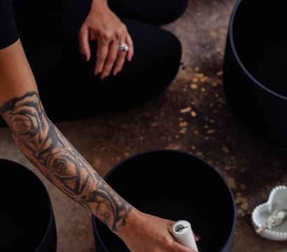A tattooed arm kneeling on a studio floor strikes a black singing bowl with a white mallet during a sound-bath meditation.